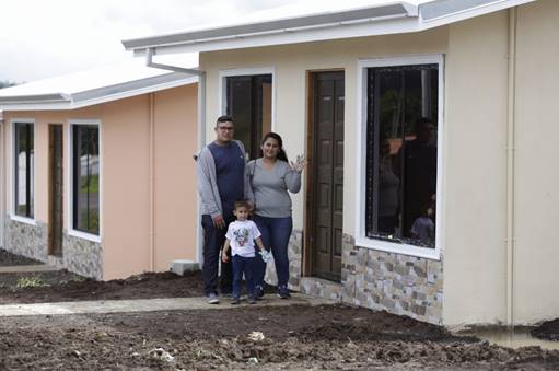 Familia constituida por dos padres jóvenes y un niño pequeño posan frente a una casa.