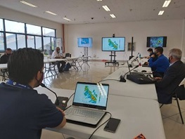Una serie de personas sentados en una mesa en forma de U dentro de una sala, con unos monitores de en la parte del frente que contienen mapas.
